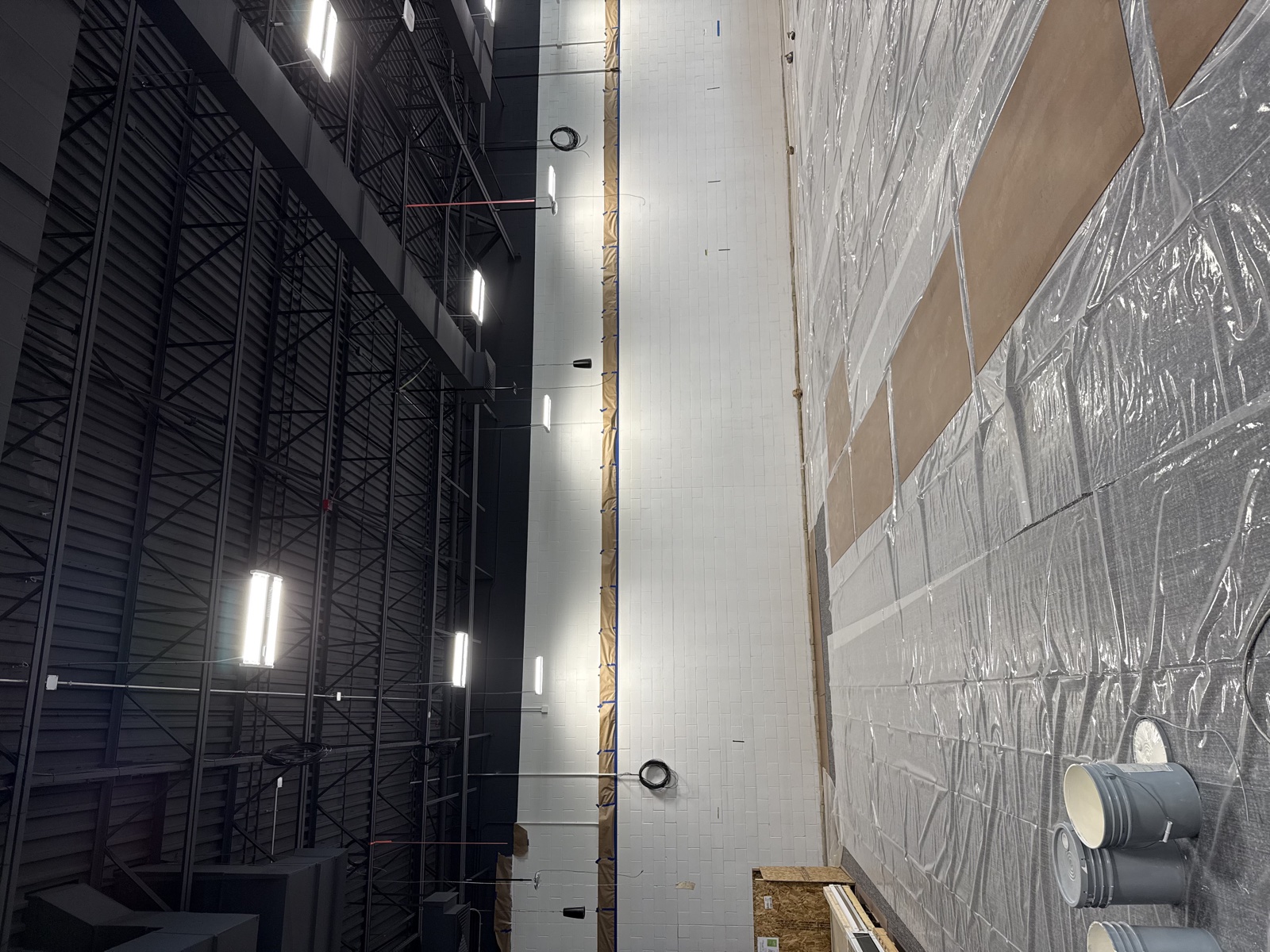 Tall industrial warehouse interior with dark walls being repainted, retail shelving wrapped in plastic sheeting, and high-ceiling fluorescent lighting overhead