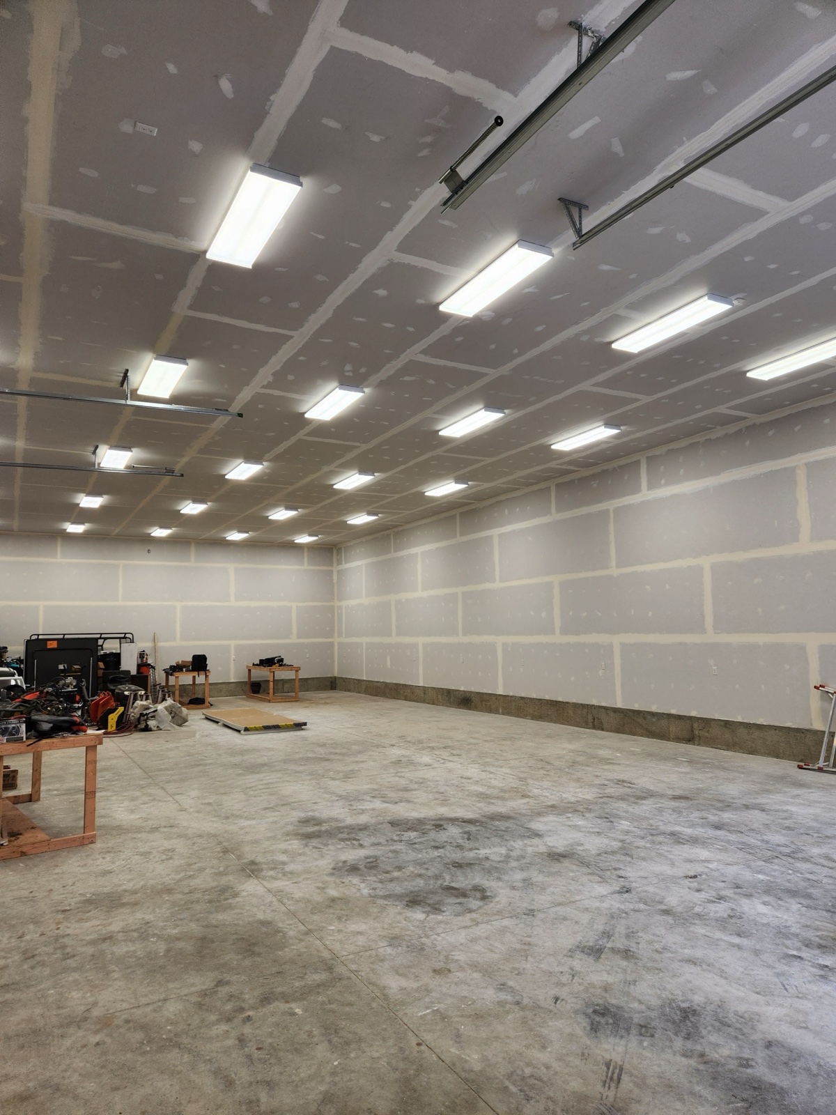 Large commercial shop interior with freshly drywalled walls and ceiling, fluorescent lighting, polished concrete floor, and lawn equipment visible in the corner