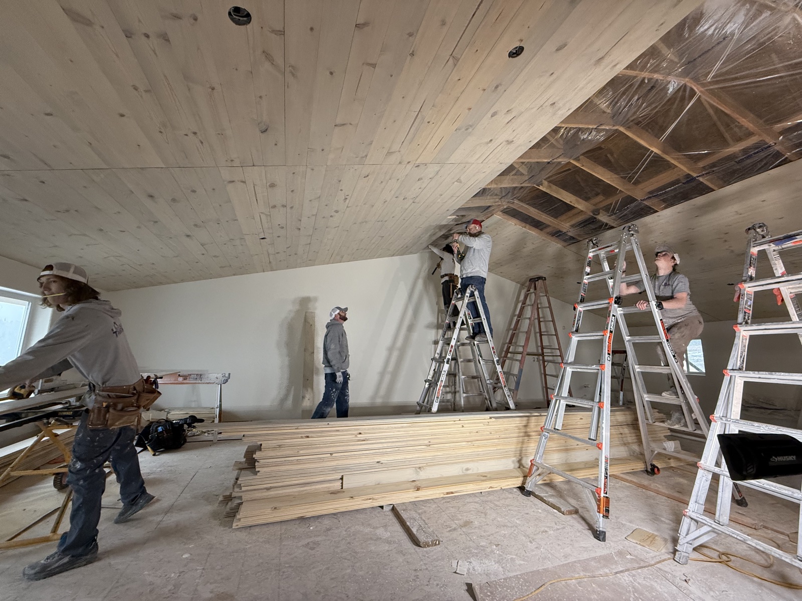 Silver Valley crew installing knotty pine tongue-and-groove boards across a tall vaulted cathedral ceiling, with stacked T&G boards on the floor and ladders set across the room