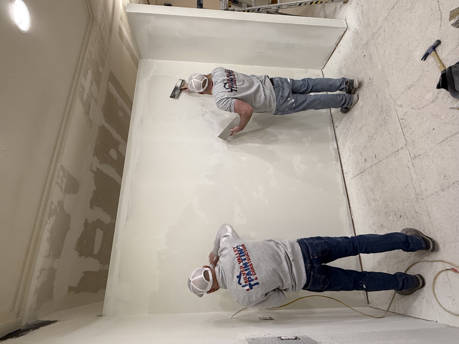 Two Silver Valley crew members in branded shirts and hard hats hand-skimming a freshly hung drywall wall during interior finish work