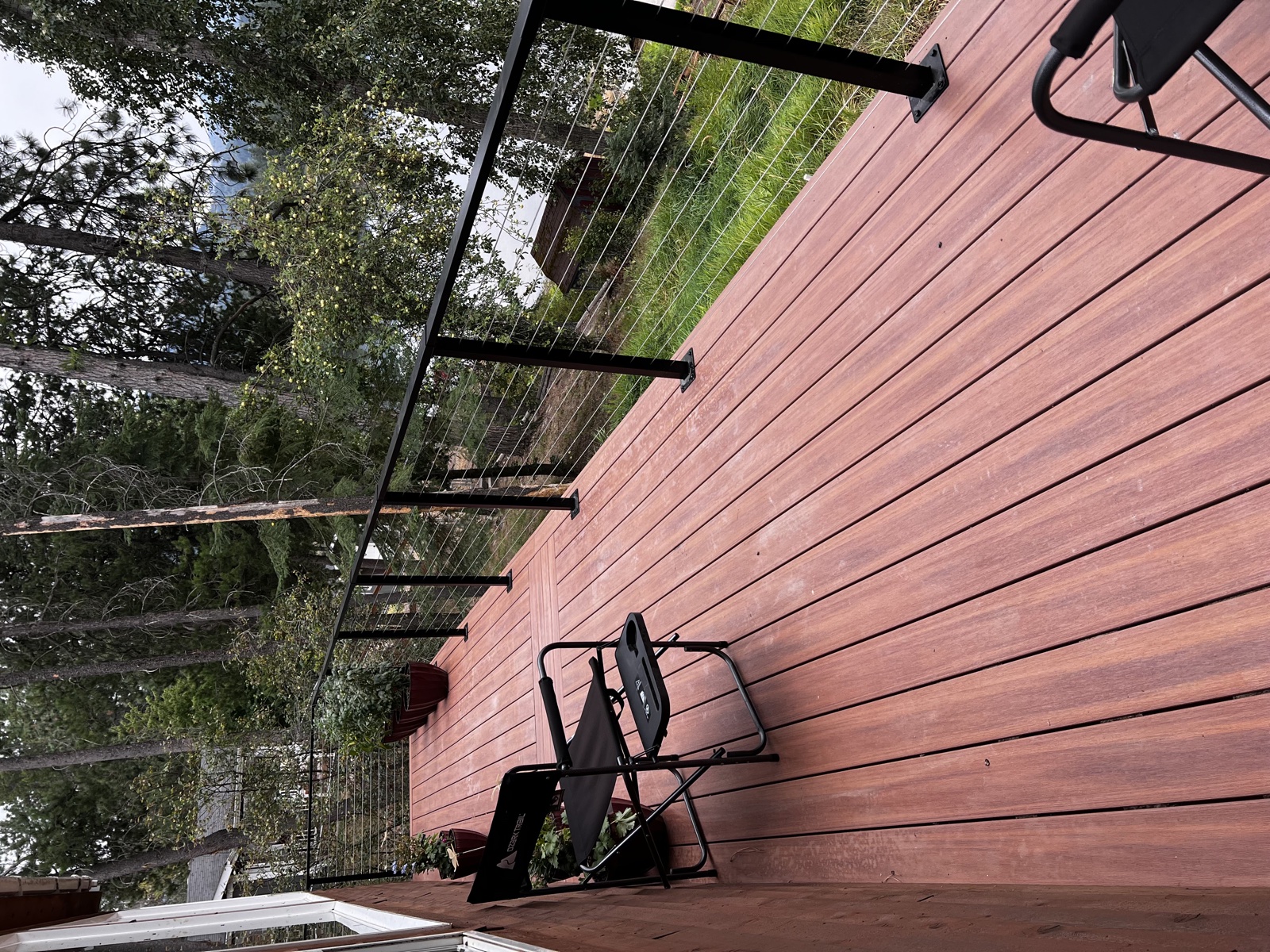 Wood deck refinished with rich red-toned stain, featuring slim black cable railing posts and a folding chair, with mature trees beyond