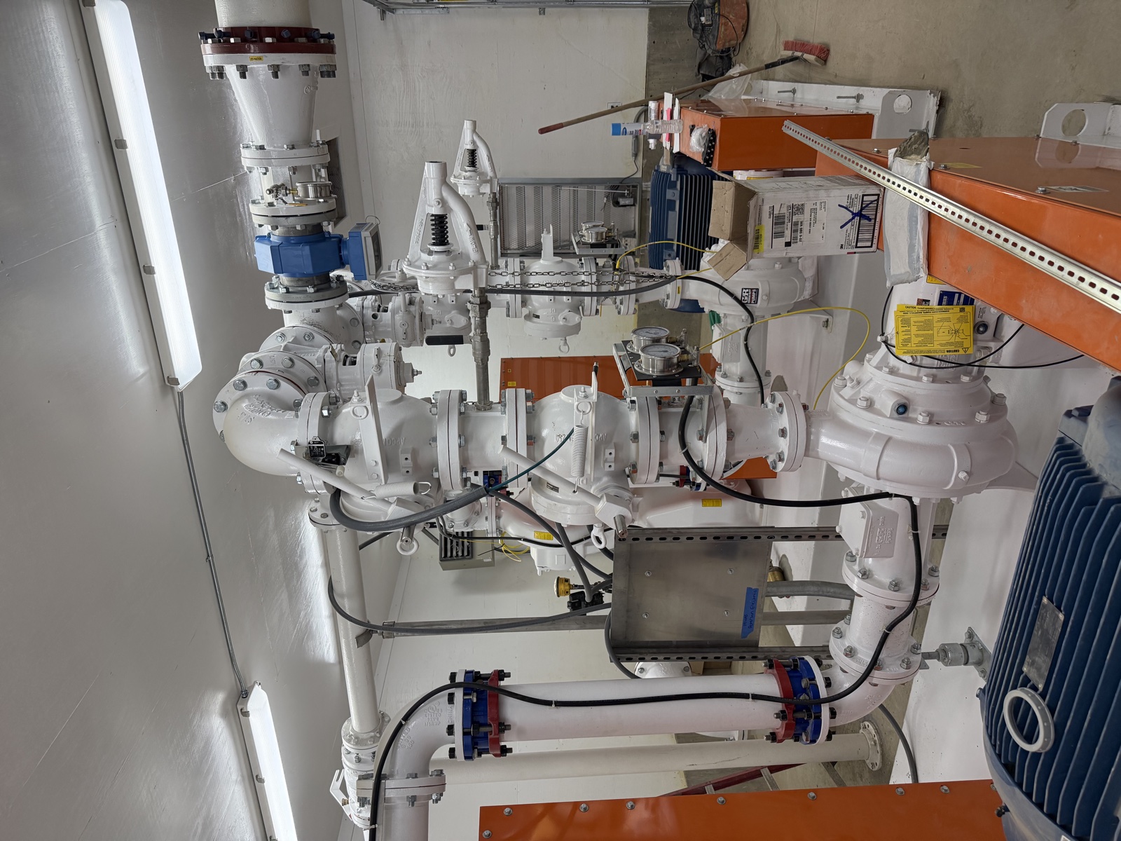 Close-up of a multi-valve manifold in a wastewater facility with white-coated piping, blue valve handles, and orange pump enclosures